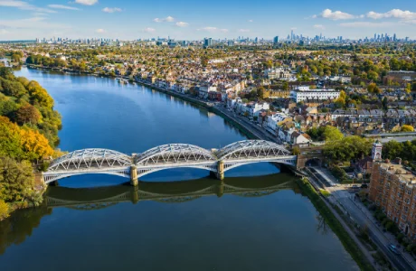 Aerial view of Barnes Bridge crossing the River Thames in Barnes, London, with surrounding riverside homes and the London skyline in the distance.